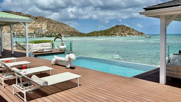 Terraza de madera con sillones de descanso y una piscina con vistas al mar azul cristalino, con montañas al fondo.