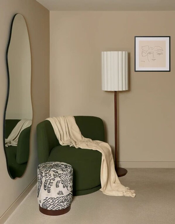 A room with a green armchair, a floor lamp, a large mirror, a framed artwork, and a patterned ottoman.
