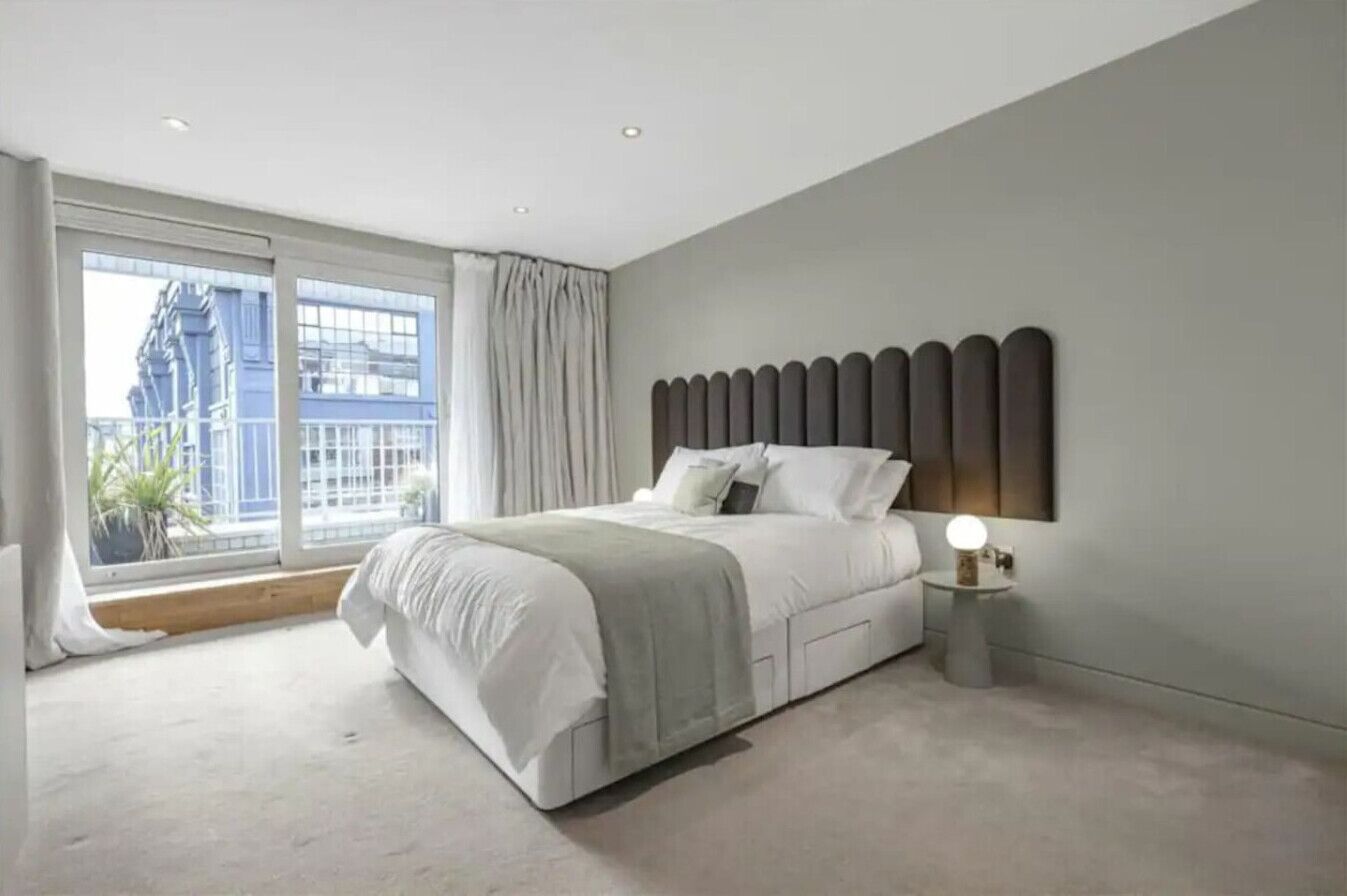 A modern bedroom with a large bed, a nightstand with a lamp, a sliding glass door to the outside, and a city view.