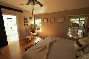 A bedroom with a large bed, a ceiling fan, a window with a view of greenery, and a door leading to a bathroom.