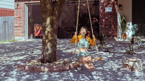 A woman sitting on a swing in a garden area with a brick house in the background.