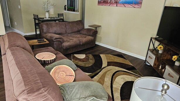 A living room with a brown sofa, a coffee table, and a TV stand with a flat-screen TV. There is a painting on the wall and a window with a view of the outdoors.