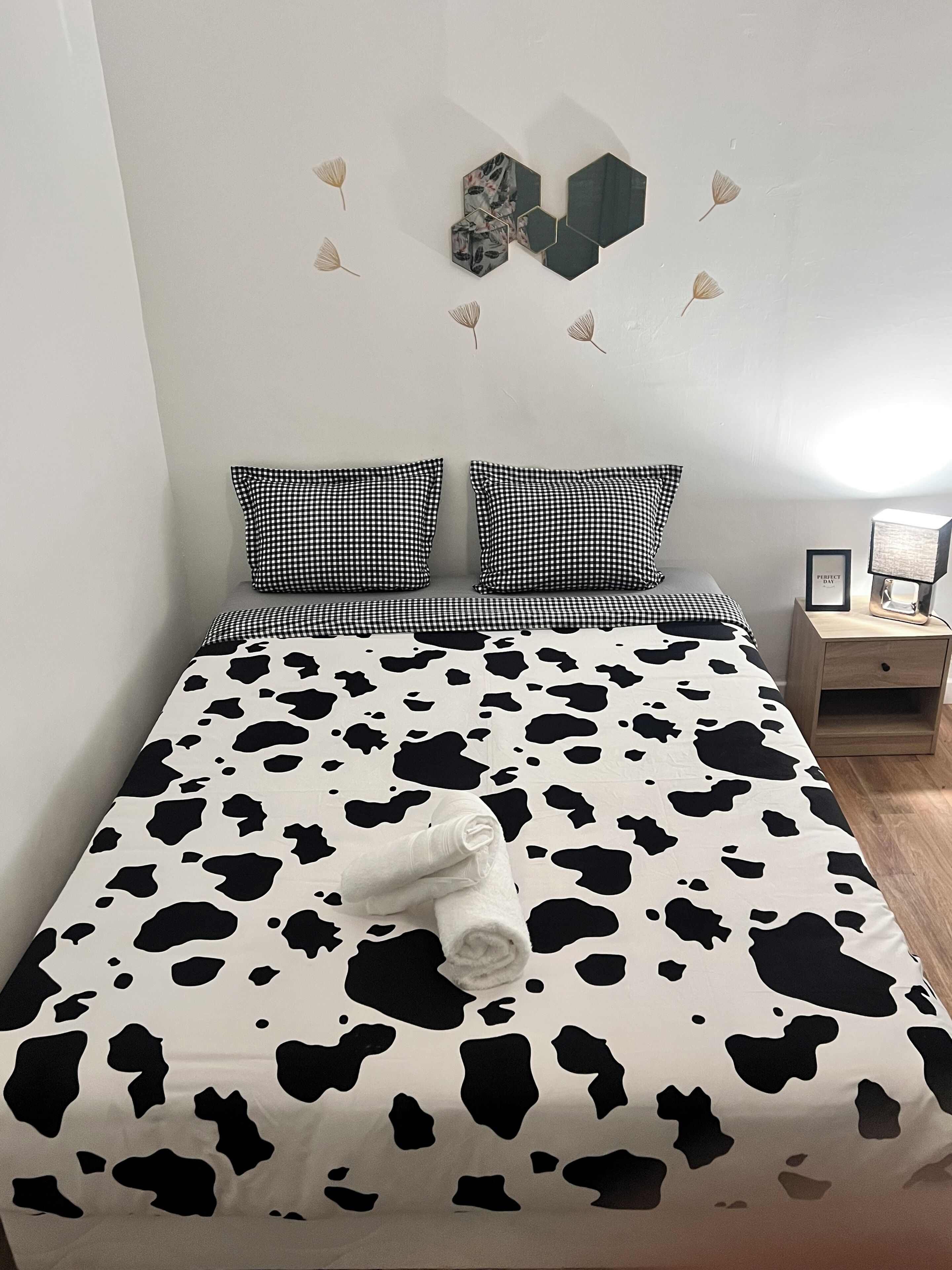 En säng med ett sängöverkast med komönstrat mönster, två rutiga kuddar och en hoprullad handduk på sängen. Bredvid sängen står ett nattduksbord med ett inramat foto och en lampa.