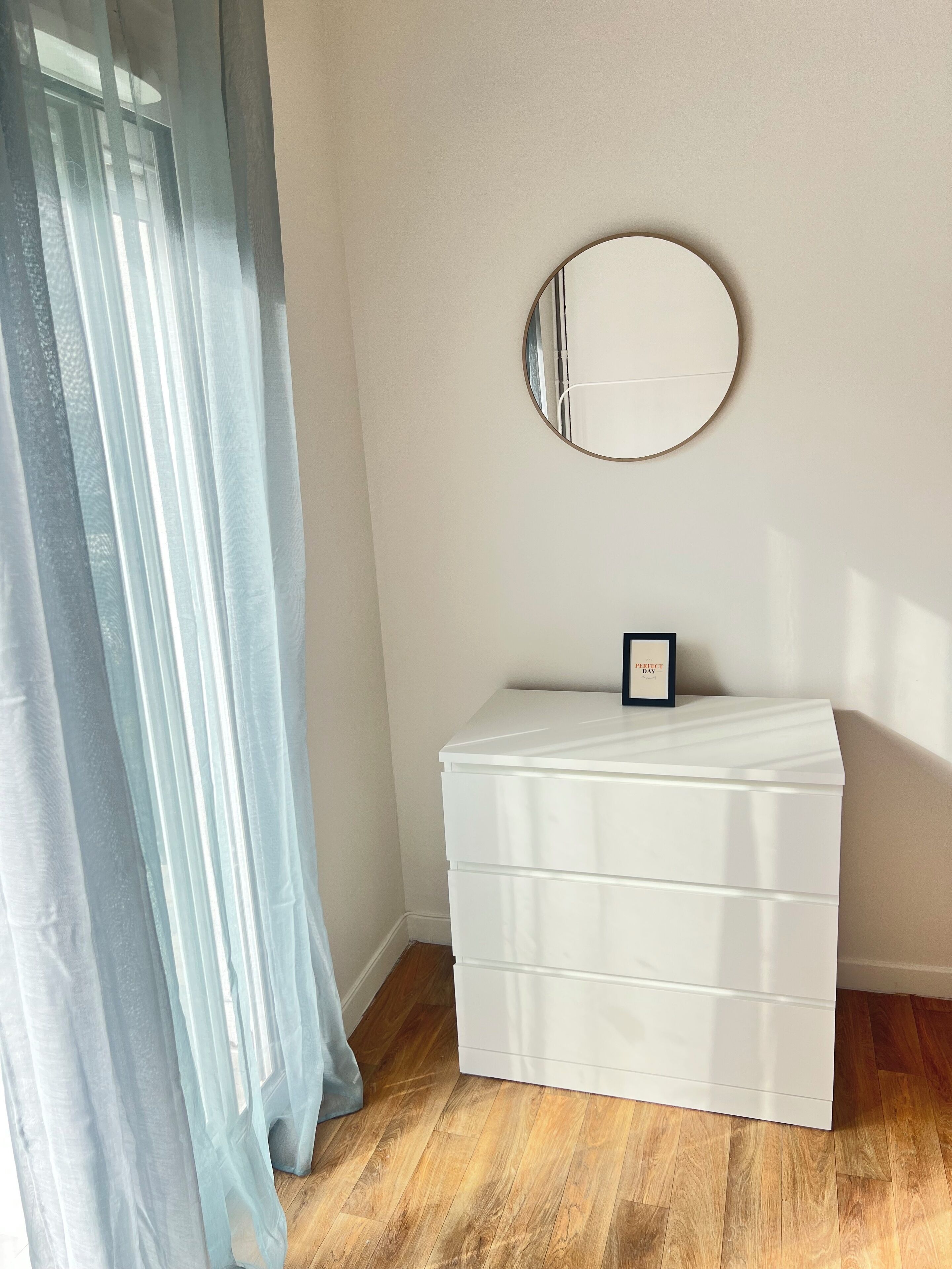 Ein Zimmer mit einer weißen Kommode, einem runden Spiegel an der Wand und durchsichtigen Vorhängen.