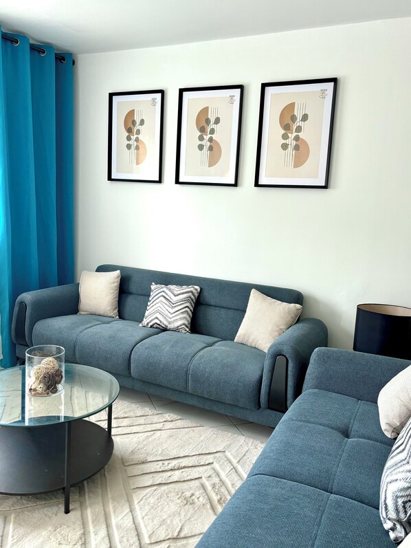 Una sala de estar con un sofá azul, una mesa de centro de vidrio y tres cuadros abstractos en la pared.