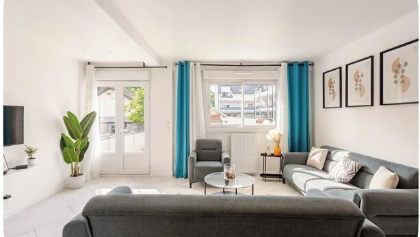 A modern living room with a grey sofa, a small round coffee table, a grey armchair, and a large window with blue curtains.