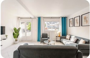 Una sala de estar moderna con un sofá gris, una mesita redonda pequeña, un sillón gris y un ventanal grande con cortinas azules.