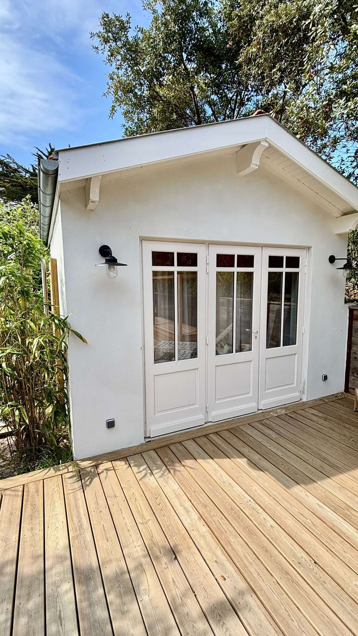 Un bâtiment blanc doté d’une terrasse en bois, de deux portes avec des panneaux de verre et d’une petite structure adjacente surmontée d’une cheminée.