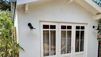 Un bâtiment blanc doté d’une terrasse en bois, de deux portes avec des panneaux de verre et d’une petite structure adjacente surmontée d’une cheminée.