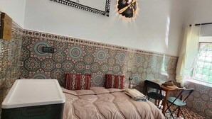 Una stanza con pareti in piastrelle decorate, un letto con copripiumino color beige, un tavolino con sedie e una finestra vista verde.