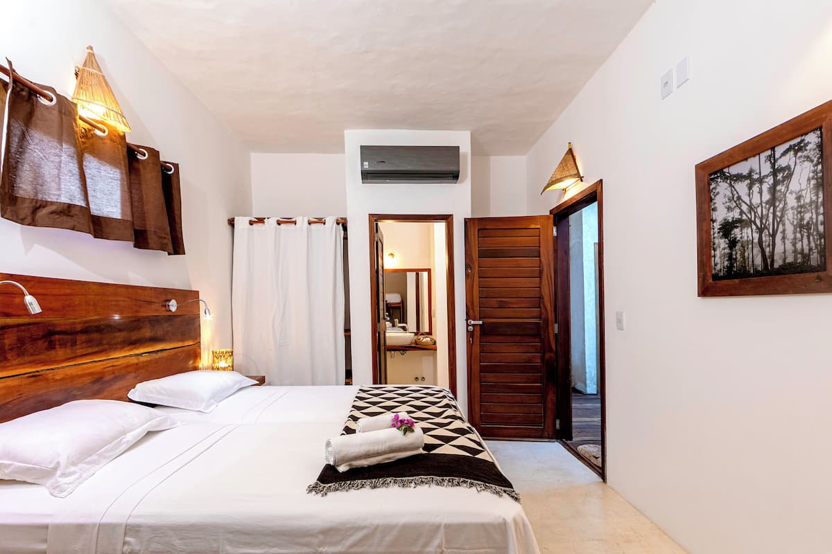 A bedroom with a wooden headboard, a bed with white linens, a geometric-patterned blanket, a wall-mounted air conditioner, and a framed picture on the wall.