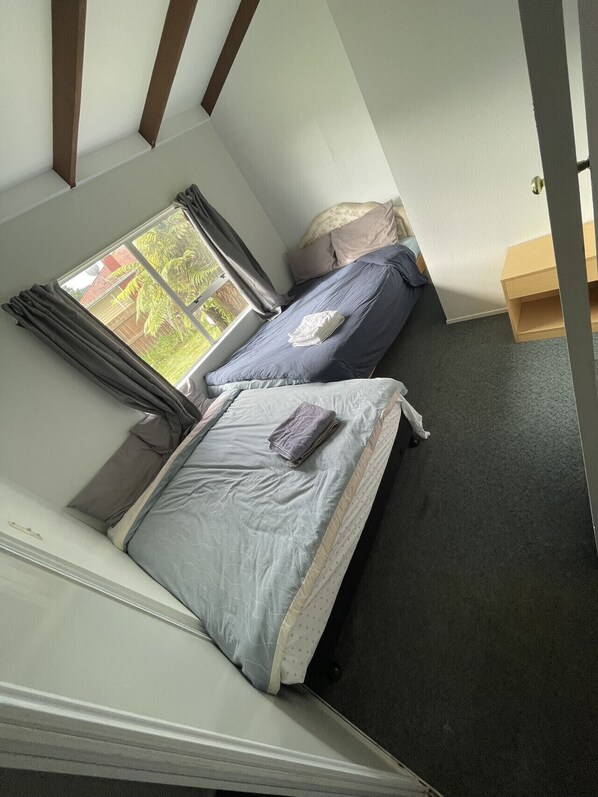 A small, compact room with two beds, a window with curtains, and a wooden shelf.