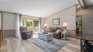 A spacious living room with a gray sofa, a gray recliner, and a white coffee table. There is a large window with curtains, a framed picture on the wall, and a small side table with a lamp. - Maple Lake Hideaway! (Longville)