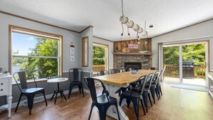 A spacious dining area with a wooden table and chairs, a stone fireplace, and large windows offering a view of the outdoors. - Maple Lake Hideaway! (Longville)