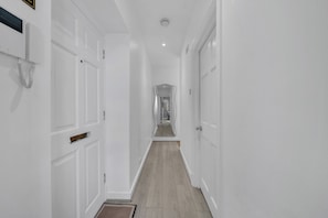 A narrow hallway with white walls and doors, a wooden floor, and a ceiling light.