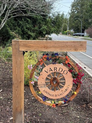 Un letrero de madera con un diseño de rueda antigua, que dice "VARDO Vintage & Valuables", rodeado de flores coloridas.