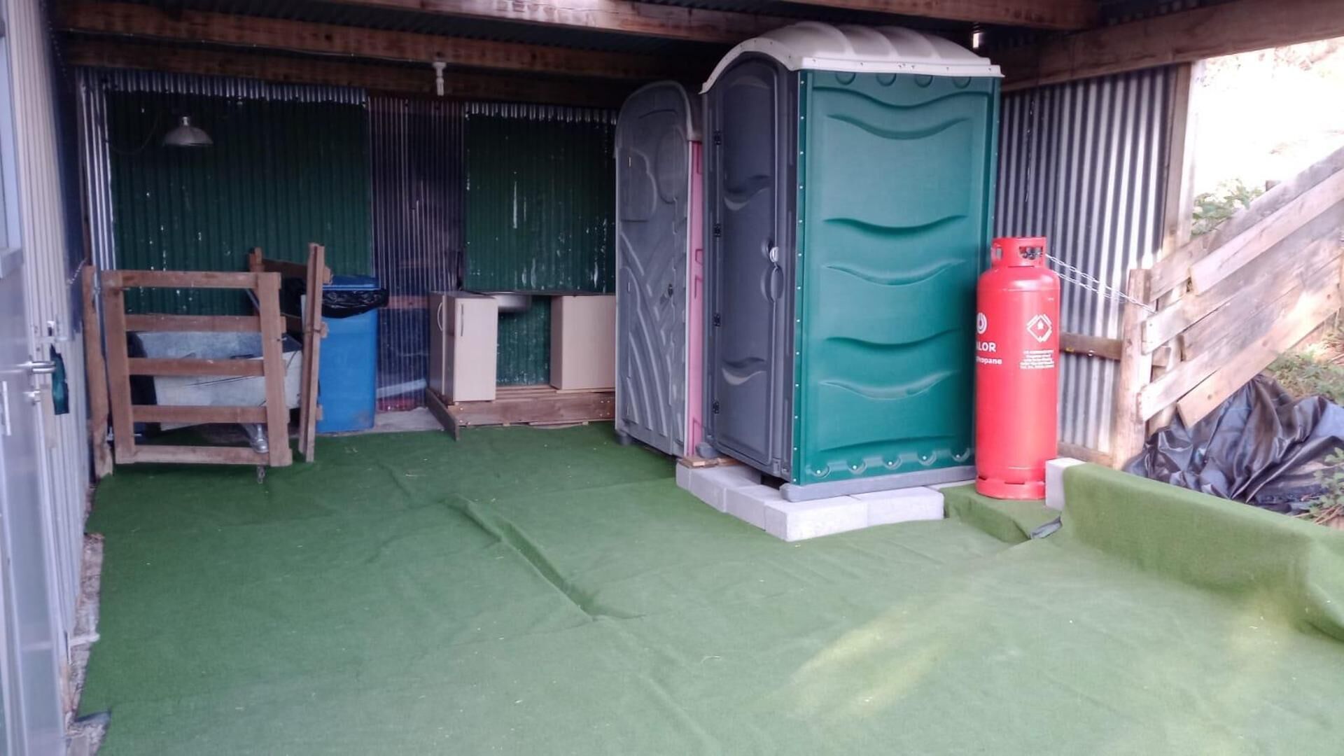 Ein kleiner, abgeschlossener Bereich mit grünem Kunstrasenfußboden, einer mobilen Toilette, einem roten Feuerlöscher und einer Holzleiter.