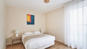 Une chambre à coucher avec un lit, des tables de chevet, une lampe, un tableau et une fenêtre avec des rideaux légers.
