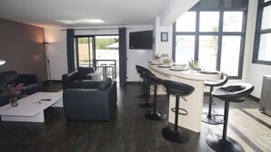 A modern living room with a sofa, coffee table, and a kitchen area with a bar stool and a TV. - Villa Luxe 8p | Piscine & Plages à Pied (Anglet)