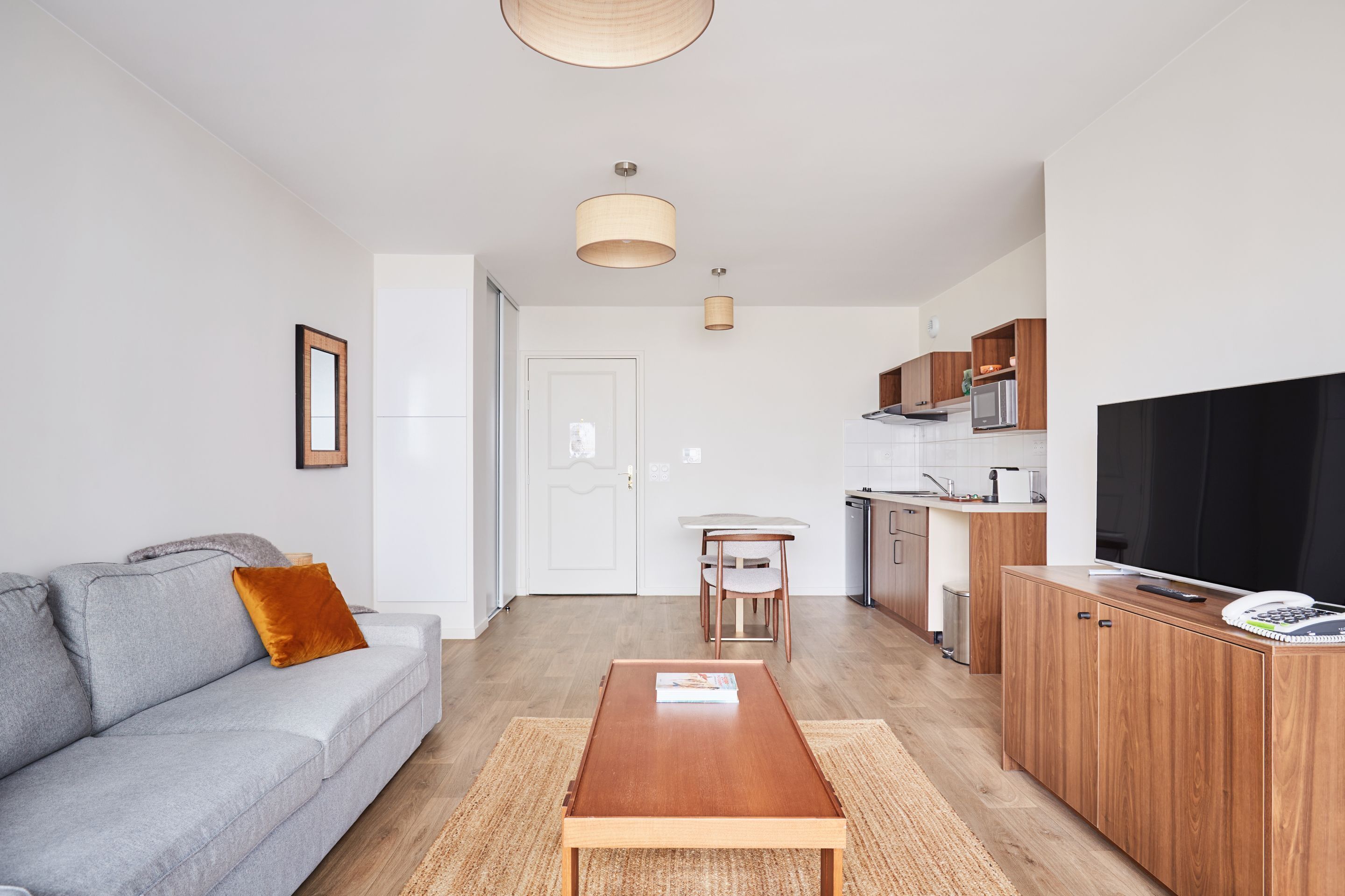 Ein modernes Wohnzimmer mit Sofa, Couchtisch und Fernseher. Es gibt eine Küchenzeile mit Schränken und einer Mikrowelle.
