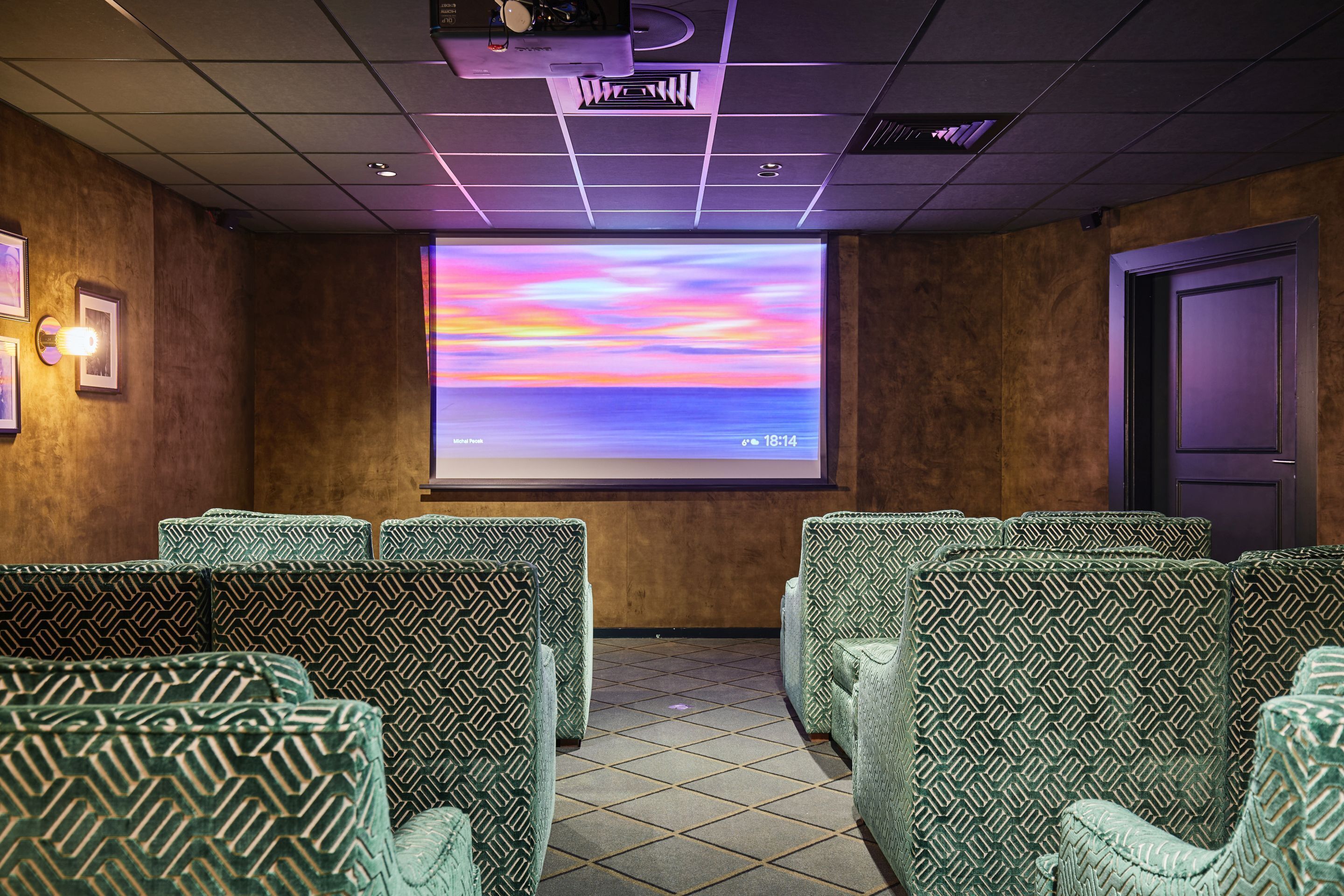 Een bioscoopzaal met gestreept groene stoelen, een groot scherm waarop een zonsondergang wordt getoond, en een projector bevestigd aan het plafond.