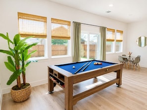 A pool table in a room with large windows, a dining area, and a potted plant.