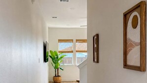A hallway with a wooden floor, a woven rug, a potted plant, and framed artwork on the walls.