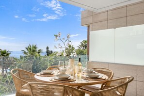 A round dining table with wicker chairs, a vase with flowers, and a bottle of wine, set against a glass balustrade overlooking a coastal view. - Sapphire | Sea view - pool - terrace (Estepona)