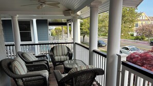 A porch with wicker furniture and a glass table, overlooking a street with parked cars and houses.