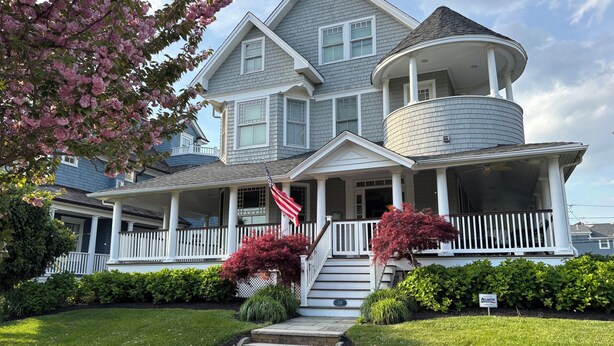 A two-story house with a wrap-around porch, a cylindrical tower, and a front yard with a lawn and shrubs.