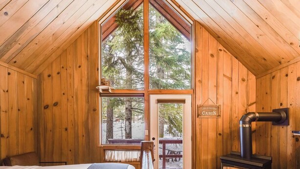 A cabin interior with a large window, a bed, a wooden ceiling, and a fireplace. - Mount Adams Cozy Cabin Retreat (Trout Lake)