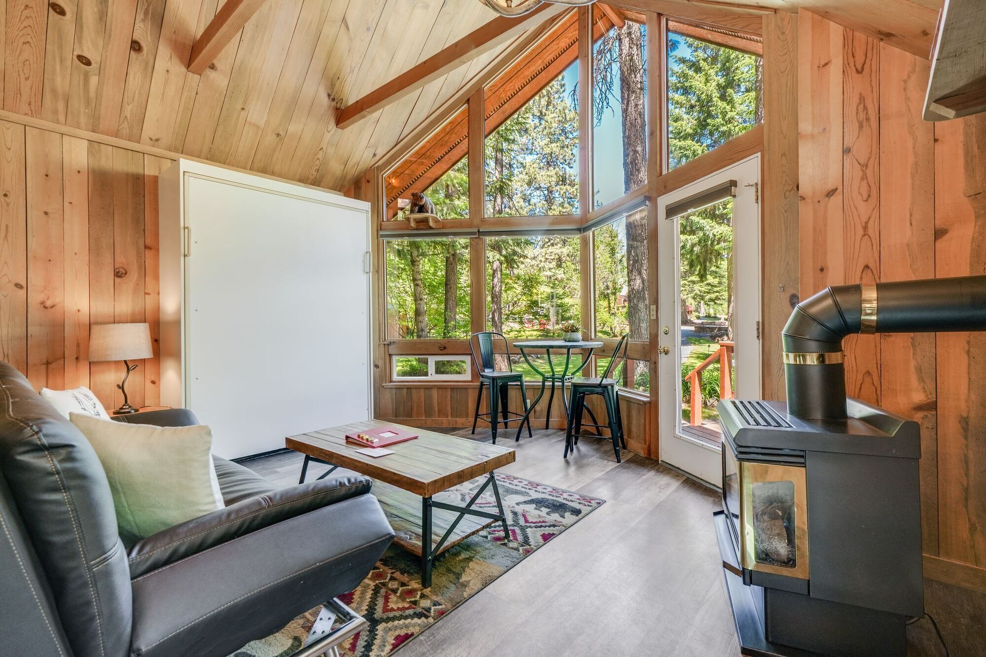 Un acogedor interior de cabaña con sofá, mesa, sillas, chimenea y ventanas amplias que ofrecen vista al bosque circundante.