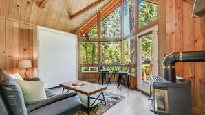 Un acogedor interior de cabaña con sofá, mesa, sillas, chimenea y ventanas amplias que ofrecen vista al bosque circundante.