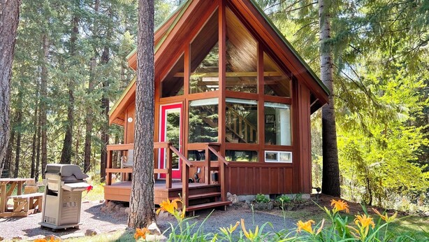 Una cabaña de madera con porche, ventanas amplias y una puerta roja, rodeada de árboles y flores.