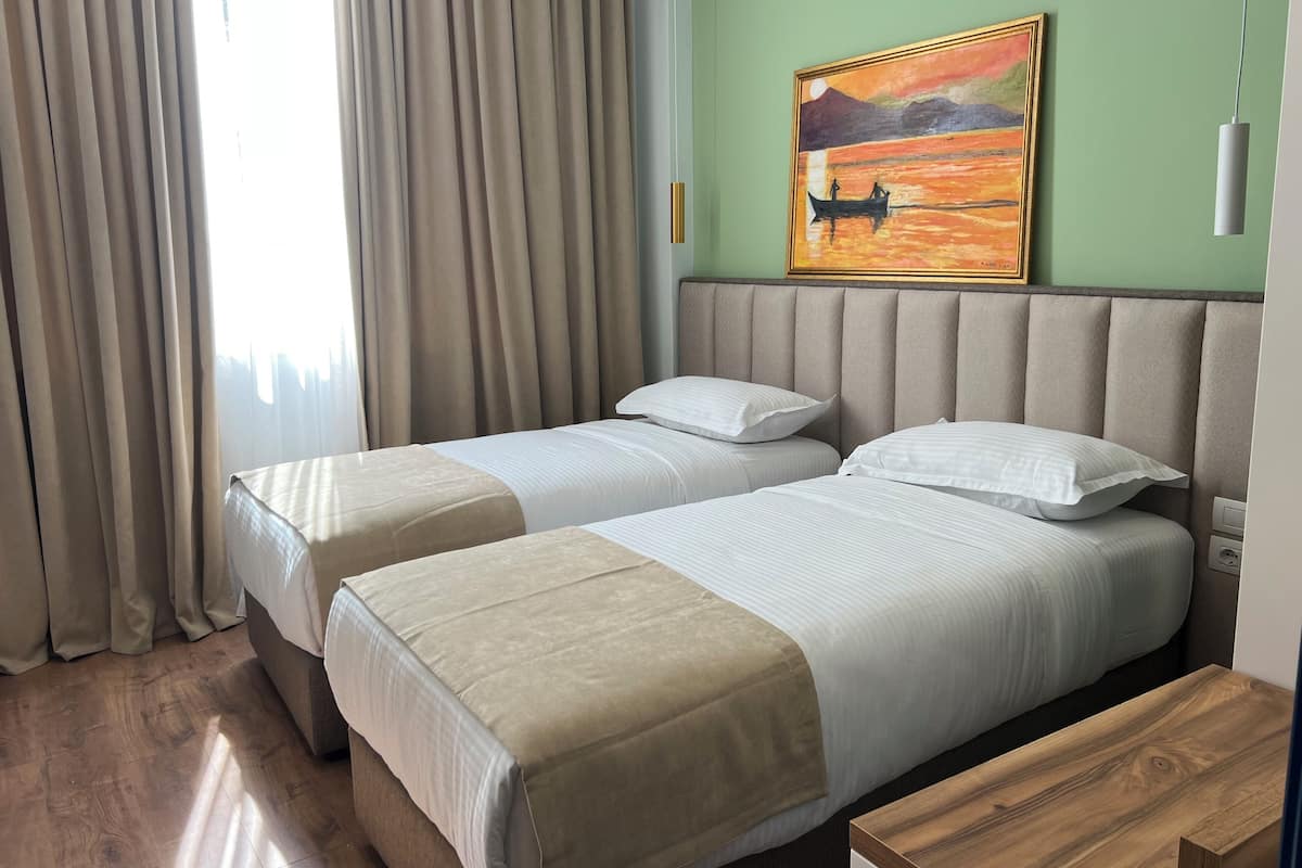 A hotel room with two beds, beige curtains, a painting of a sunset, and a wooden nightstand.