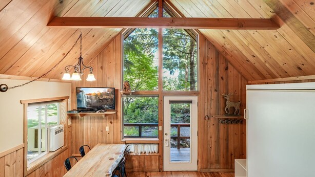 A cabin with a wooden interior, a dining area, and a large window offering a view of the forest. - Beautiful Cabin With Partial Views of Mt. Adams (Trout Lake)