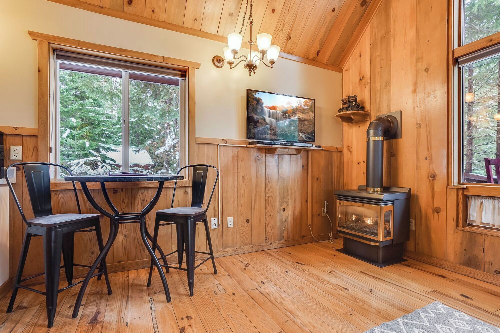 Interior acogedor de una cabaña con mesa y sillas de madera, chimenea, televisor montado en la pared y ventanas amplias que dan a vistas del bosque circundante.
