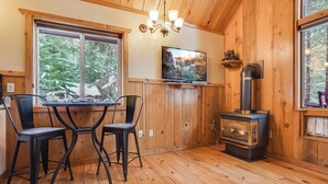 Een gezellig cabin interieur met een houten eettafel en stoelen, een open haard, een aan de muur bevestigde televisie en grote ramen met uitzicht op het omliggende bos.