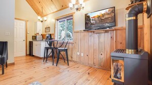 Een gezellig interieur van een chalet, met een houten plafond, een open haard, een eethoek met stoelen, een keuken met een magnetron en een tv boven de open haard.