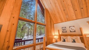 Een knus kamer in een chalet, met een groot raam, houten wanden, een bed met kussens met hertenmotieven en twee wandlampen.