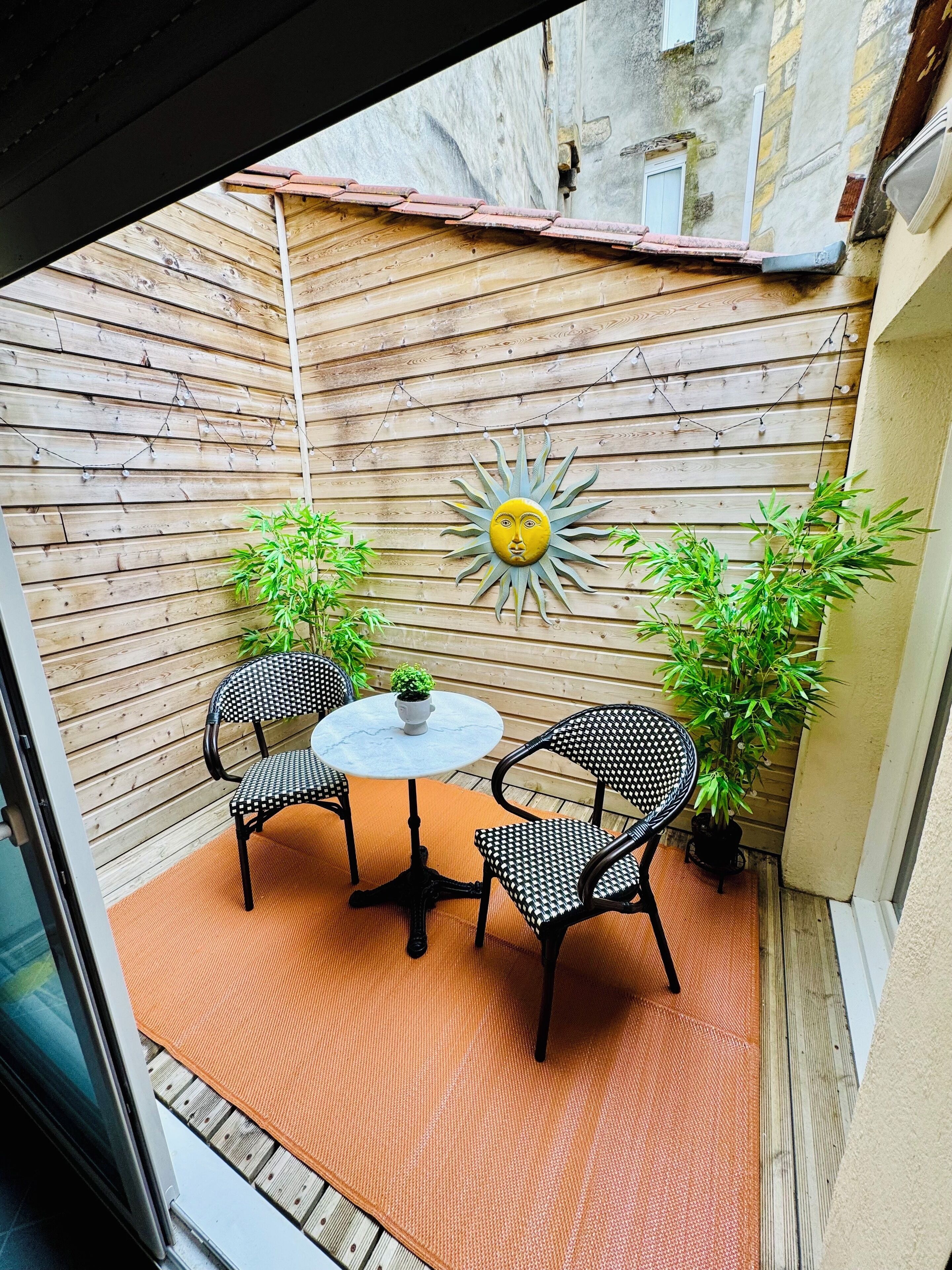 Un piccolo patio con un tavolo rotondo e due sedie, circondato da listelli di legno e piante in vaso.