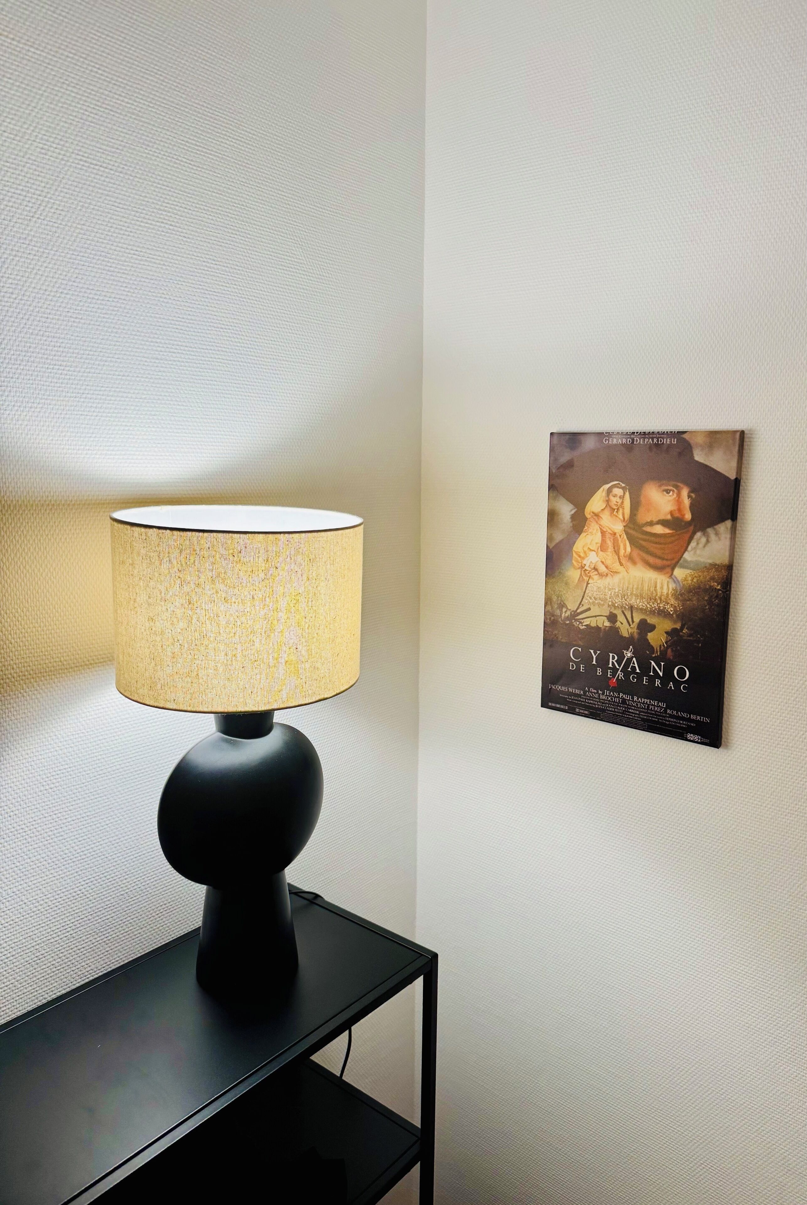 A lamp with a textured lampshade on a black table against a textured wall. A framed movie poster is visible on the wall.