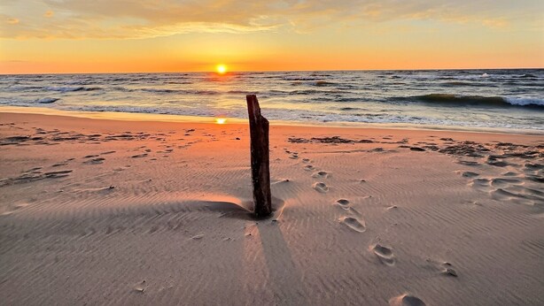 Una playa con huellas que conducen a un poste de madera, y el sol poniéndose sobre el océano.
