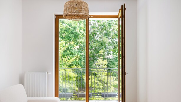 A room with a white sofa, a wooden floor, a white radiator, and a view of a balcony with greenery.