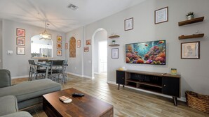 Ein modernes Wohnzimmer mit Couchtisch aus Holz, einer grauen Couch, einem Essbereich mit Stühlen und einem an der Wand befestigten Fernseher.