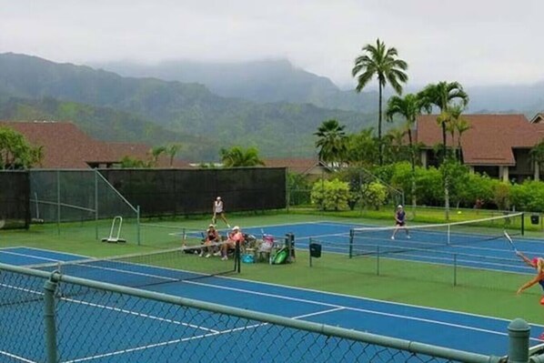 Una cancha de tenis con una red, rodeada de follaje y palmeras, con montañas al fondo.