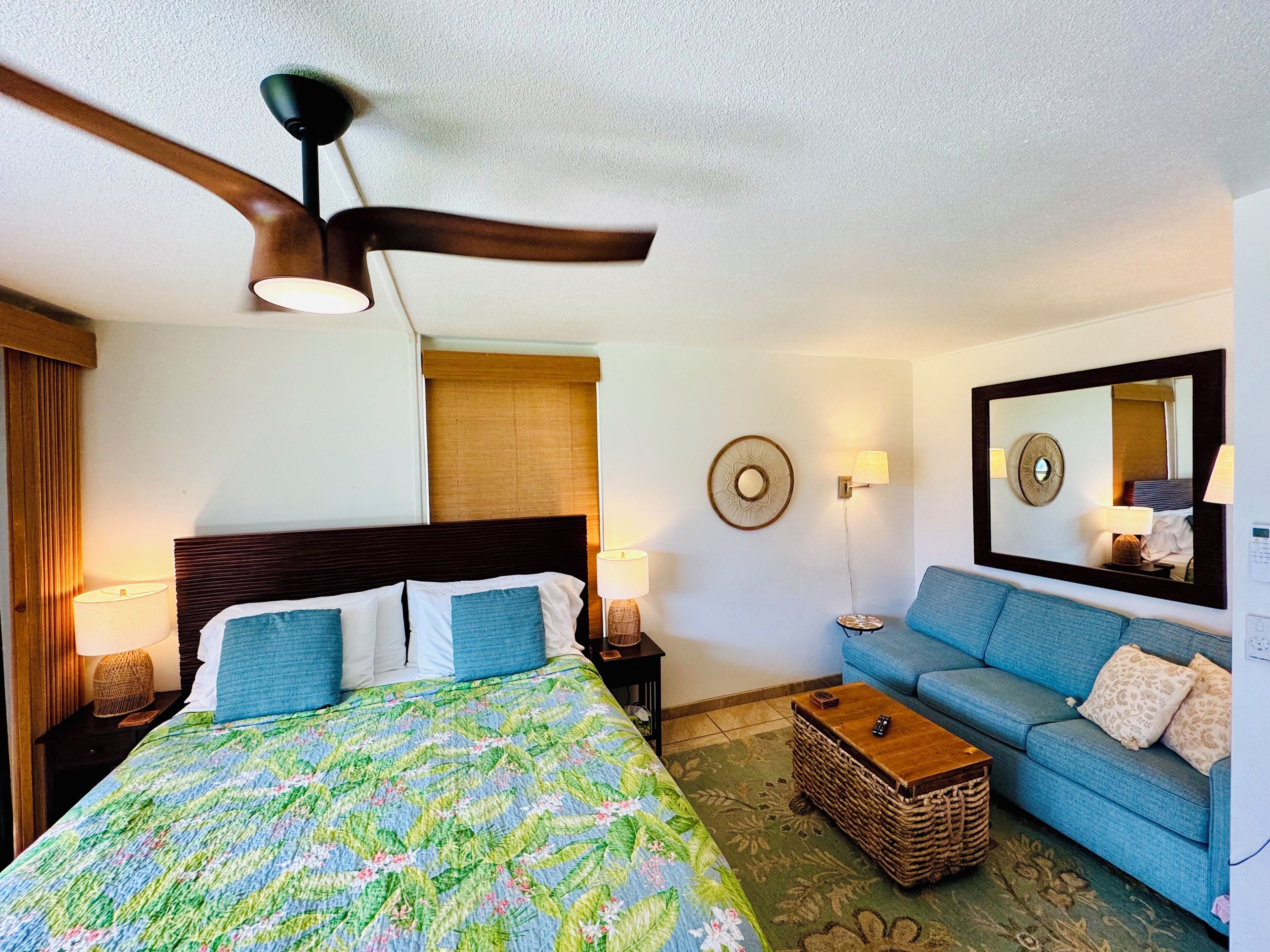 Quarto de hotel com cama, sofá azul, ventoinha no teto, espelho fixado na parede e uma pequena mesa.