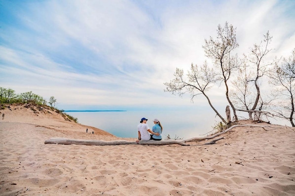 Una playa arenosa con una pareja sentada sobre un tronco, contemplando una masa de agua tranquila.