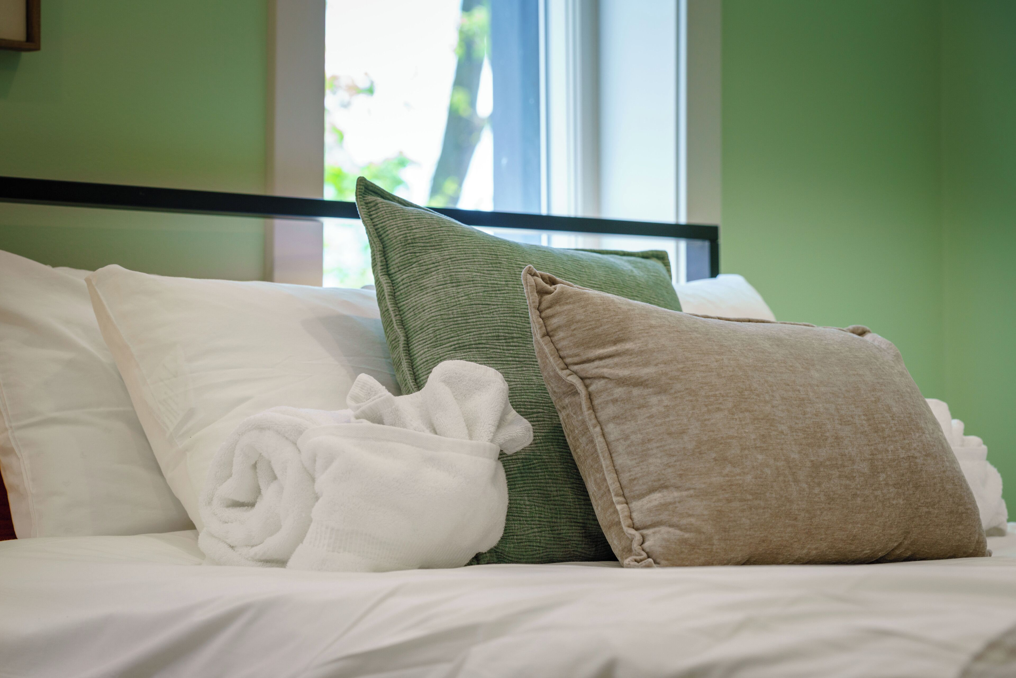 Una cama bien tendida con almohadas y una toalla enrollada, una ventana con vistas a la vegetación, y una pared verde.