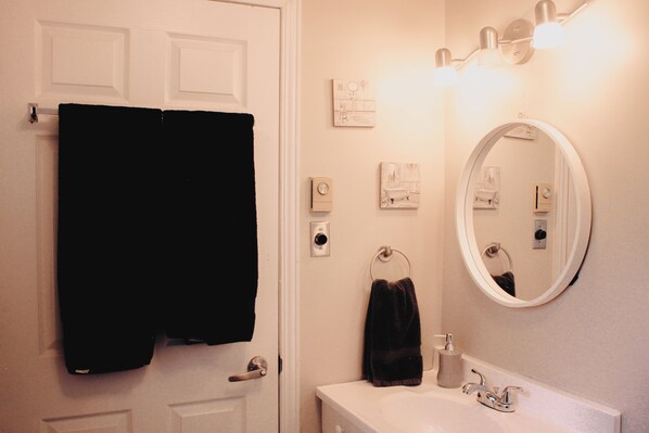 A bathroom with a white door, a black towel hanging on the door, a white sink with a mirror above it, and a towel ring on the wall. - Kingston Varty Lake Cabin: Hot Tub •Fire Pit •BBQ (Yarker)